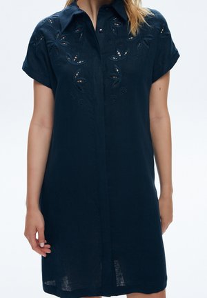 Woman wearing a dark navy short-sleeve shirt dress with floral cutout embroidery on the chest and shoulders.