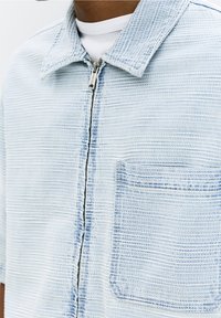 Světlá modrá džínová bunda s texturovaným povrchem, zapínáním na zip a jednou náprsní kapsou. Má límec a krátké rukávy.