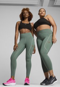Reggiseni sportivi neri, leggings verde oliva con accenti e logo bianchi, indossati con scarpe da ginnastica rosa e nere. Texture liscia, design aderente.