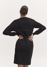 Abito nero lavorato a maglia con maniche lunghe, vita stretta e orlo a ginocchio. Tessuto morbido, design minimalista e nessun motivo o accessorio visibile.
