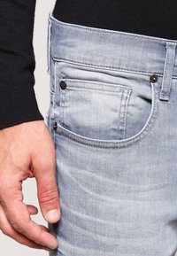 Jeans de denim azul claro con un ajuste slim, que cuentan con un bolsillo trasero con detalles de costura y remaches metálicos, mostrados junto a un suéter negro.