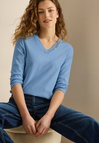 Hellblaue langärmelige V-Ausschnitt-Pullover mit geripptem Ausschnitt, kombiniert mit dunkelblauen weit geschnittenen Jeans, sitzt auf einer hellen Oberfläche.