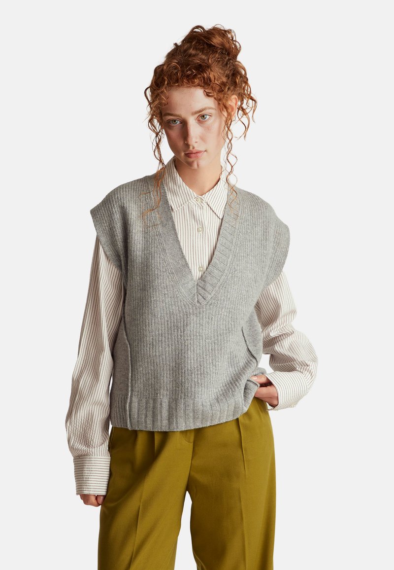 Grauer ärmelloser Pullover mit V-Ausschnitt und gerippter Textur, kombiniert mit einem weißen gestreiften Langarmshirt und olivgrünen Hosen.