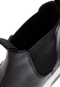 Primer plano de un zapato negro de piel tipo slipper con un panel lateral elástico texturizado y una lengüeta en el talón, mostrando la abertura interior y el borde de la suela blanca.