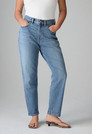Person iført lyseblå jeans med højtaljede, med hænderne i lommerne og thongsandaler med lille hæl på en ensfarvet grå baggrund.
