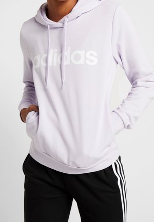 Person iført en lyse lilla Adidas hættetrøje med frontlomme og sorte bukser med hvide sidelængder.