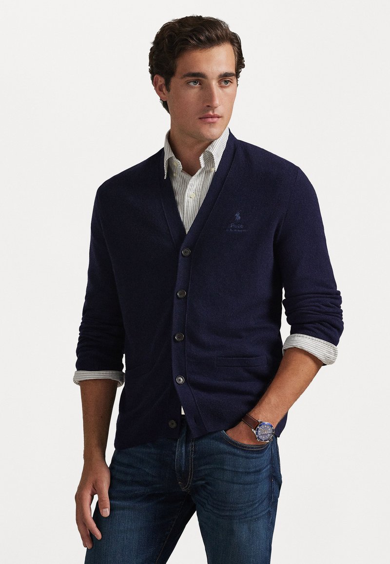 Cárdigan azul marino con escote en V, cinco botones y dos bolsillos. Combinado con una camisa de collar a rayas y jeans azules.