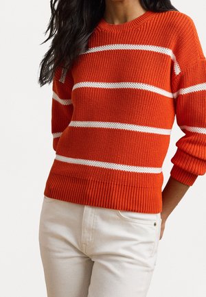 Femme portant un pull en tricot orange vif avec des rayures horizontales blanches et un pantalon clair, debout avec une main derrière le dos.