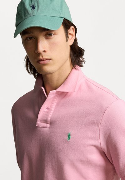 Camisa polo rosa con cuello y una tapeta de un solo botón, con un logo bordado en verde. Acompañada de una gorra verde claro.