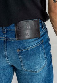 Siniset farkut, joissa on teksturoitu nahkapaikka, johon on kirjoitettu "GIANNI KAVANAGH" takana. Klassiset taskut ja istuva muotoilu.