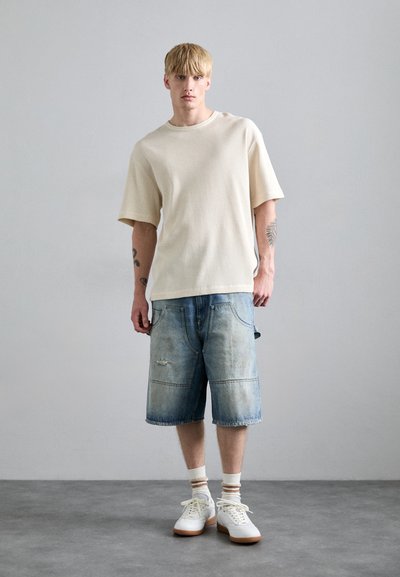 Beige teksturert t-skjorte med korte ermer, kombinert med lyseblå denimshorts med synlig søm og slitte detaljer. Hvite sneakers med brune såler.