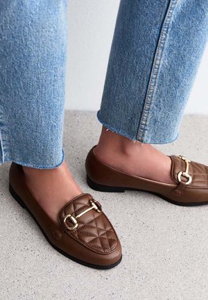 Brown leather loafers with a quilted design and gold-tone hardware accent. Worn with cropped blue denim jeans. Flat sole, rounded toe.