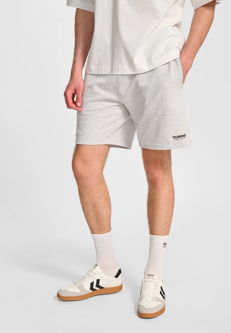 Lichtgrijze katoenen shorts met een zwart "hummel" logo. Gecombineerd met witte sportsokken en witte sneakers met zwarte strepen en gumzolen.
