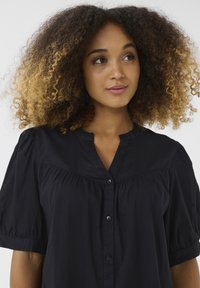 Camicia di cotone nera con collo a V, maniche a sbuffo e chiusura con bottoni, caratterizzata da delicate arricciature al petto e una texture liscia.