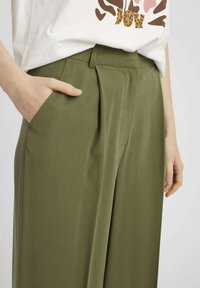 Pantalons vert olive à coupe décontractée, dotés de poches latérales et d'un design plissé. Fabriqués en tissu lisse avec une texture uniforme.