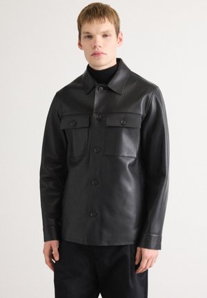 Young person wearing a black leather button-up jacket with chest pockets over a black turtleneck, standing against a plain light background.