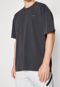 Tmavě šedé bavlněné tričko s kulatým výstřihem, oversized střihem, krátkými rukávy a jemným detailem loga na hrudi. Měkká textilie s vybledlým vzhledem.