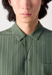 Green collared shirt with vertical stripes, featuring a textured fabric, button closure, and a front pocket on the left.