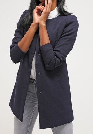 Femme portant un long blazer bleu marine foncé sur une chemise blanche et un jean gris clair, les mains touchant son visage et son menton sur un fond uni.