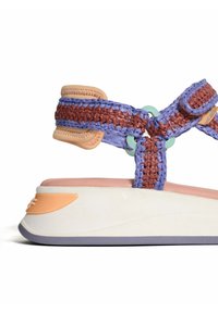 Schoen met banden met een gevlochten paarse en bruine textiele bovenkant, een zachte roze voetbed en een lichte witte zool met oranje en grijze accenten.