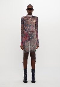 Robe longue transparente à manches longues avec un motif floral multicolore, un col haut et des fentes sur les côtés, associée à des bottines noires et des lunettes de soleil.