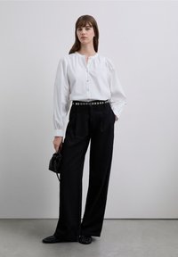 Camisa blanca con mangas abullonadas y tapeta de botones, combinada con pantalones negros de talle alto y cinturón con tachuelas. Zapatos negros y bolso pequeño.
