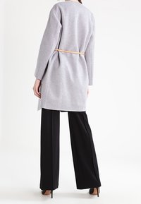Vrouw in een lange grijze gewatteerde jas en zwarte wijde broek, staand met de rug naar de camera op een witte achtergrond.