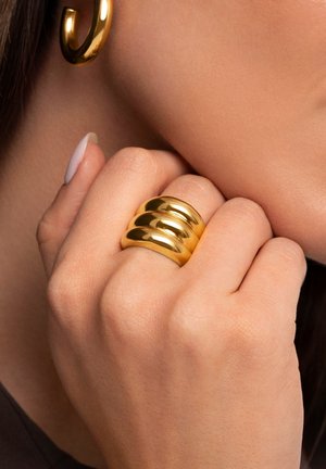 Anello in tono oro con tre bande lisce e arrotondate impilate insieme, che riflettono la luce con una finitura lucida. Indossato su una mano parzialmente visibile.