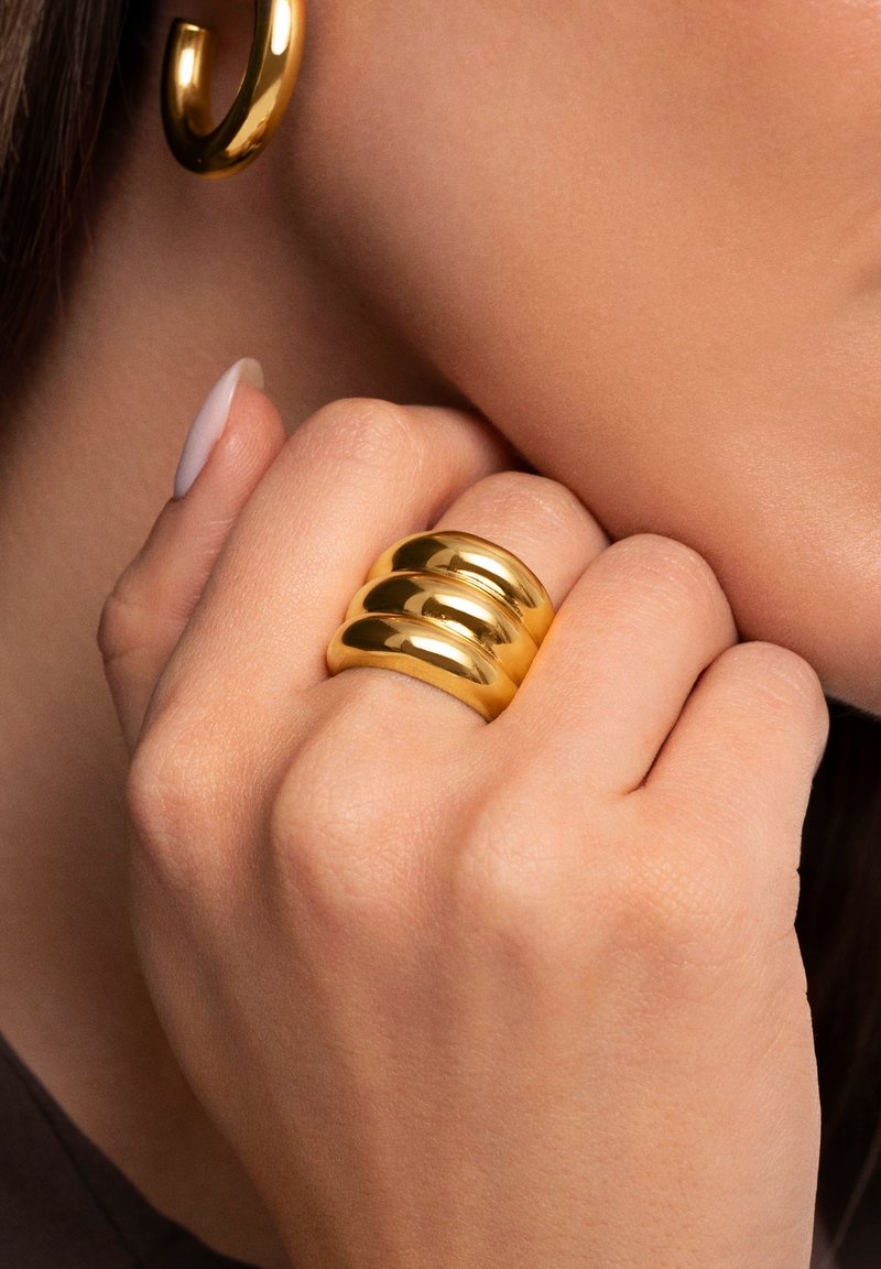 Anello in tono oro con tre bande lisce e arrotondate impilate insieme, che riflettono la luce con una finitura lucida. Indossato su una mano parzialmente visibile.