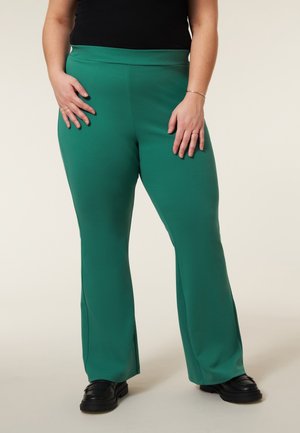 Groene, uitlopende broeken met een gladde textuur, hoge taille en zijsplitten. Draag ze met zwarte schoenen, die een effen kleur hebben zonder patronen.