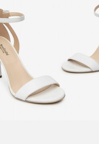 Sandali alti bianchi con un design elegante. Presentano una ampia fascia anteriore e una cinghia alla caviglia, realizzati in materiale sintetico liscio.