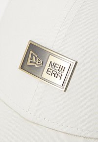 Hvit baseballcap med teksturert stoff, med et rektangulært metallmerke med en hevet logo med påskriften "NEW ERA."