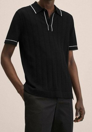 Mężczyzna ubrany w czarną koszulkę polo z krótkim rękawem, z białym wykończeniem na kołnierzyku i rękawach, w ciemnych spodniach, stojący na jednolitym tle.