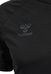 Camiseta deportiva negra con detalle texturizado en los hombros, que presenta un sutil logo gris "hummel" y un diseño de abeja en la zona del pecho.