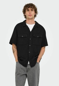 Sort kortærmet skjorte med knapper og to forlommer, tekstureret stof og spredt krave, båret over en hvid t-shirt og grå jeans.