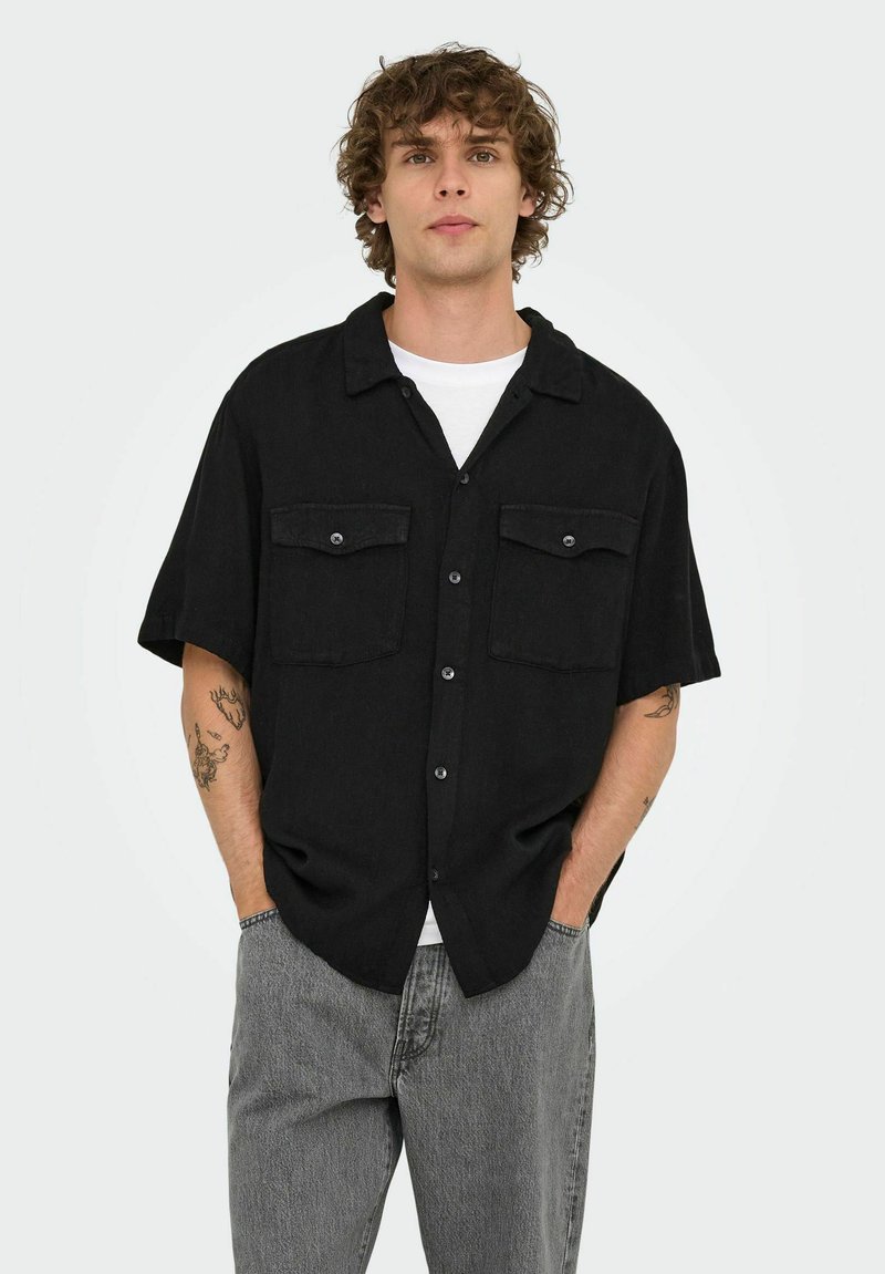 Sort kortærmet skjorte med knapper og to forlommer, tekstureret stof og spredt krave, båret over en hvid t-shirt og grå jeans.
