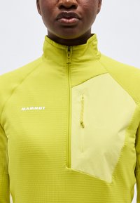 Κοντινό πλάνο ενός ατόμου που φοράει φωτεινό κίτρινο μπουφάν Mammut με φερμουάρ και τσέπη στο στήθος με φερμουάρ.