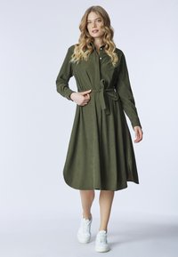 Abito verde oliva con maniche lunghe, bottoni sul davanti e vita legata, design fluido a trapezio e orlo all'altezza del ginocchio. Abbinato a sneakers bianche.