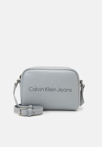 Šedá kabelka přes rameno z umělé kůže s vyraženým vzorem "CK" a značkováním "Calvin Klein Jeans". Obsahuje nastavitelný popruh a obdélníkový tvar.