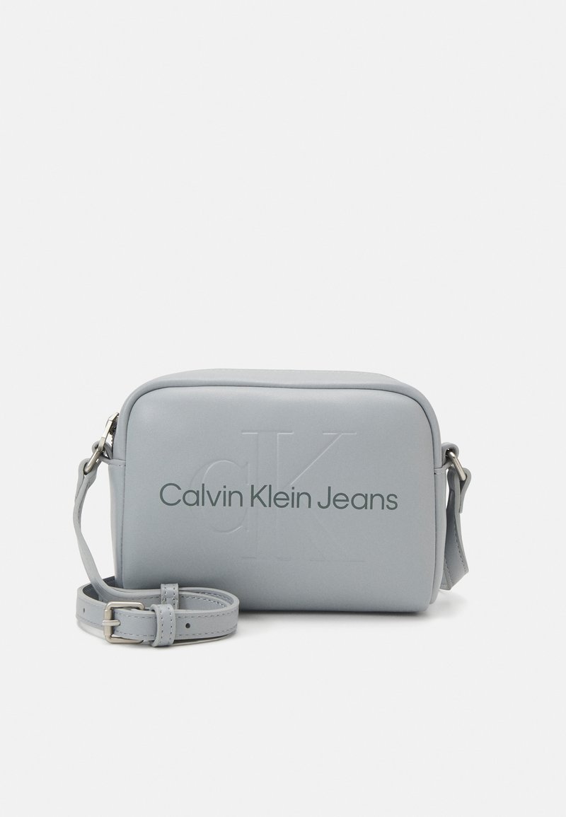 Šedá kabelka přes rameno z umělé kůže s vyraženým vzorem "CK" a značkováním "Calvin Klein Jeans". Obsahuje nastavitelný popruh a obdélníkový tvar.