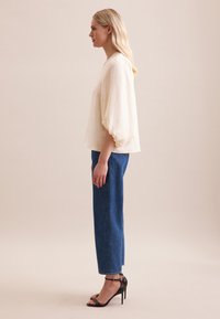 Blusa in lino color crema con maniche a sbuffo, abbinata a jeans blu a vita alta. La modella indossa dei sandali neri con tacchi alti.