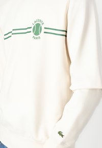 Cream sweatshirt featuring green stripes and a tennis ball graphic with "LACOSTE PARIS." Includes a green embroidered crocodile logo on the sleeve.