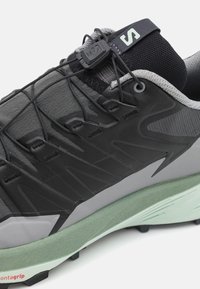 Scarpa da corsa con una tomaia in mesh nero e grigio, sovrapposizione in gomma testurizzata, lacci sicuri e un intersuola verde chiaro per un'ammortizzazione ottimale.