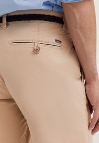 Beige Hosen aus leichtem Stoff, mit einer geknöpften Gesäßtasche, einem geflochtenen schwarzen Gürtel und einem Etikett am Bund.