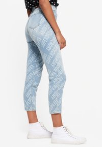 Jean bleu clair au design ajusté, avec un motif texte "SUPERDRY" blanc sur toute la surface. Accompagné de baskets blanches montantes.