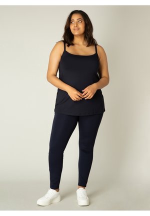 Navy Tanktop mit dünnen Trägern, kombiniert mit navyfarbigen Leggings. Weicher, dehnbarer Stoff mit glatter Textur. Weiße Sneakers. Einfaches, lässiges Design.