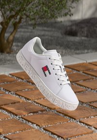 Baskets blanches Tommy Jeans avec patch logo et semelle embossée, exposées au-dessus d'un revêtement extérieur en carreaux de bois et gravier.