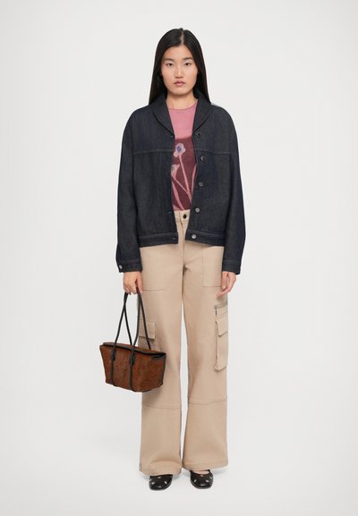 Frau in einer dunklen Jeansjacke über einem rosa blumigen Pullover, beigen weiten Cargo-Hosen, schwarzen Schuhen, die eine braune Handtasche mit schwarzen Trägern hält.