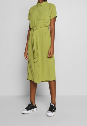 Femme portant une robe boutonnée longueur genou de couleur vert olive avec manches courtes et ceinture nouée à la taille, associée à des baskets noires basses.