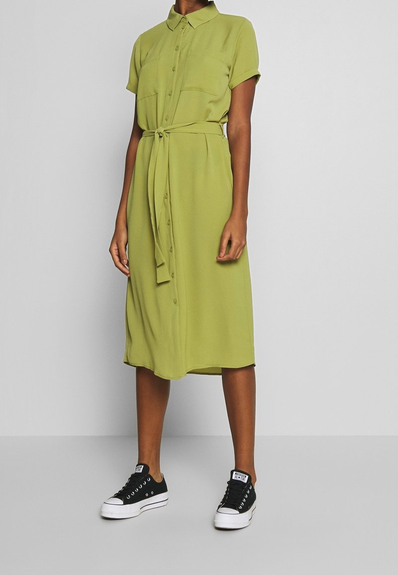 Femme portant une robe boutonnée longueur genou de couleur vert olive avec manches courtes et ceinture nouée à la taille, associée à des baskets noires basses.